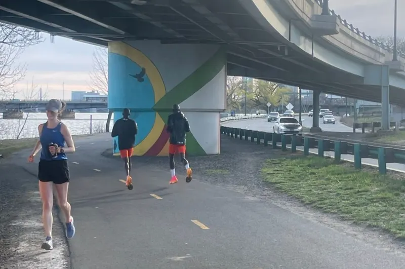 Three runners train on a riverside path beneath a bridge
